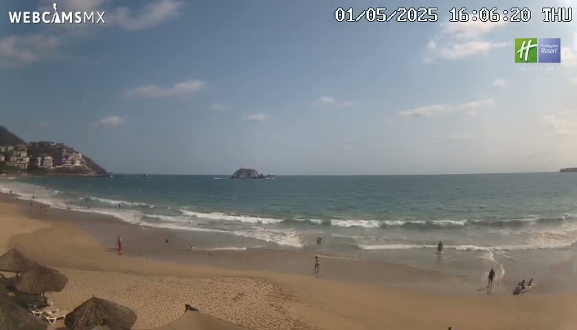 A sandy beach with gentle waves laps the shore under a mostly sunny sky, with a few clouds, and hills visible in the distance.