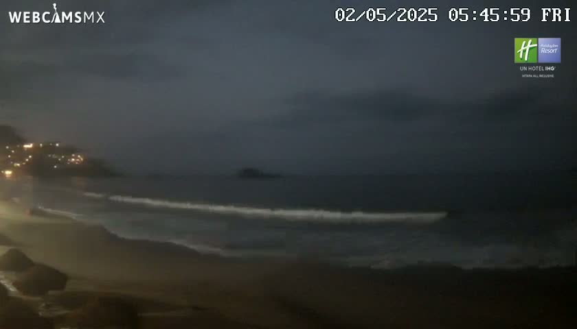A dark, overcast night scene shows a beach with gently lapping waves and a distant, dimly lit town.