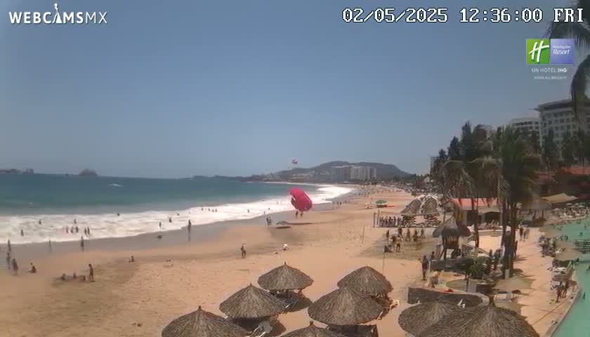 A sandy beach with a few people on it and calm ocean waves under a clear blue sky, with parasailers overhead and palm trees and hotels in the background.