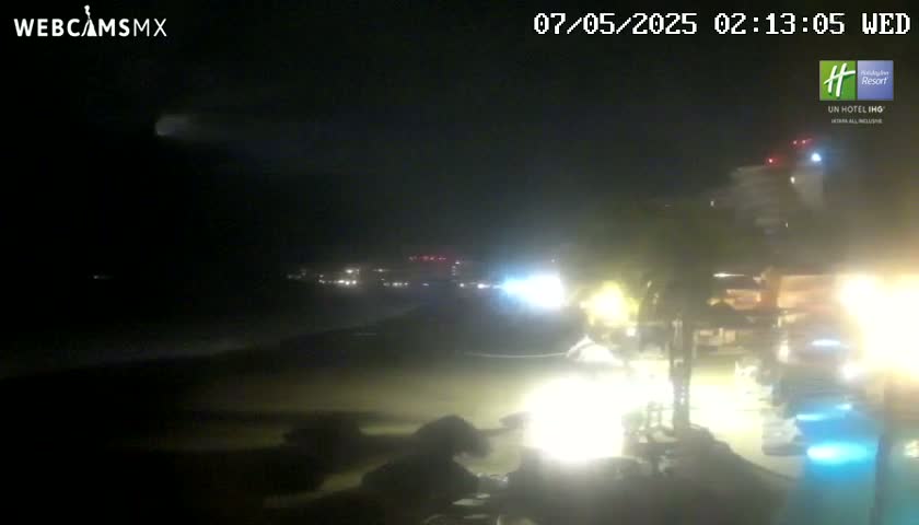 A nighttime view of a beach at night, showing a dark ocean and dimly lit beachfront buildings with lights visible on the beach and in the water.