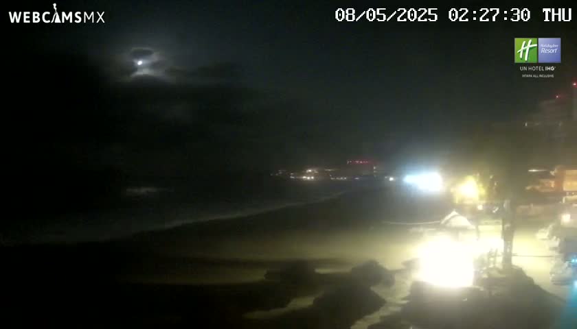 A nighttime beach scene under a partly cloudy moonlit sky shows dark ocean waves lapping the shore near dimly lit buildings.