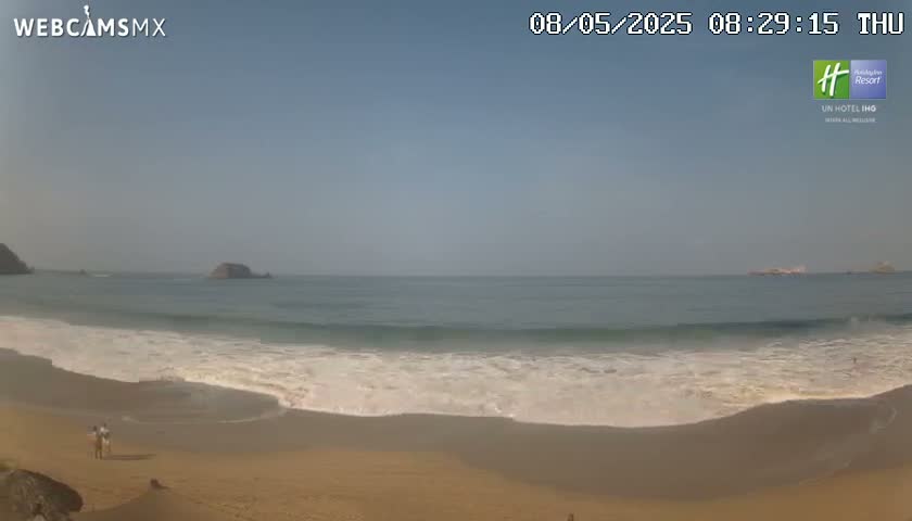 A sandy beach with gentle waves rolling onto the shore under a clear, sunny sky; two people are visible walking on the beach in the distance.