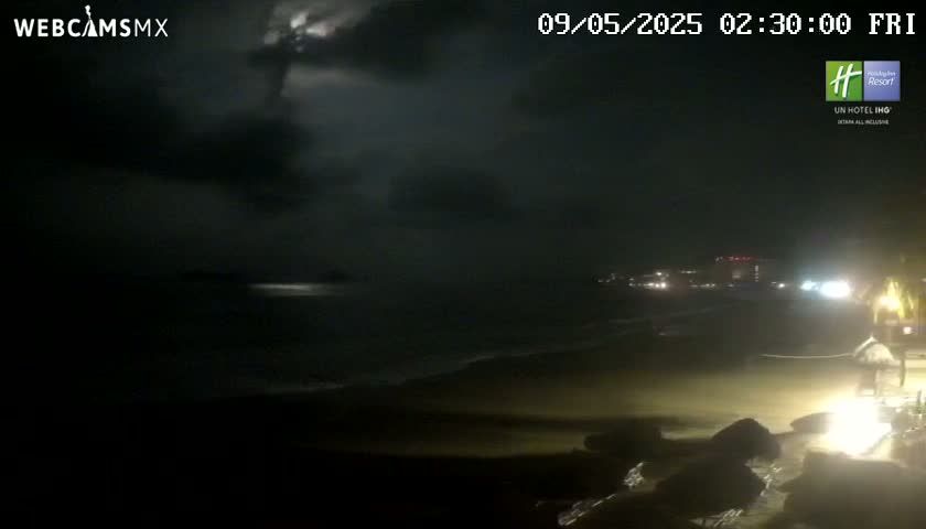 A dark, nighttime beach scene shows a cloudy sky with lightning in the distance, and dimly lit buildings and beach features along the shore.