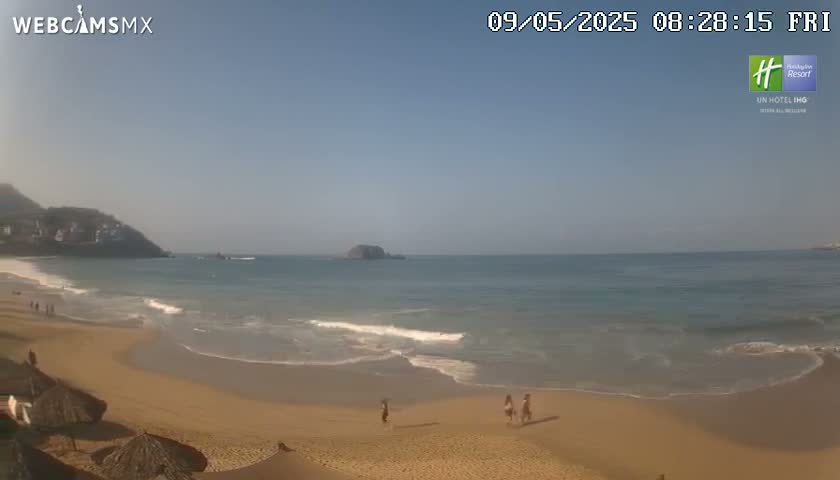 A sandy beach with small waves and a few people on it, backed by a hill and calm ocean under a clear, sunny sky.