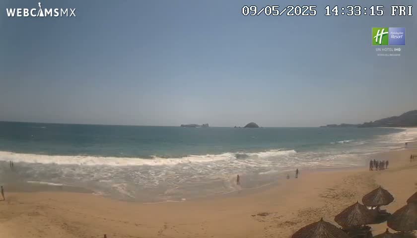 A sandy beach with small waves and a few people on it stretches to a calm ocean under a clear, sunny sky.