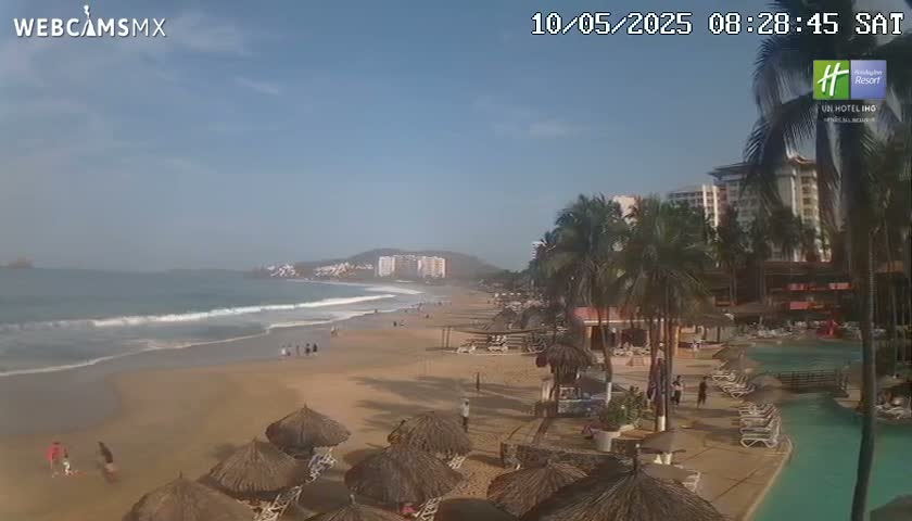 A sandy beach with calm waves, palm trees, resort buildings, and thatched umbrellas under a mostly clear sky.