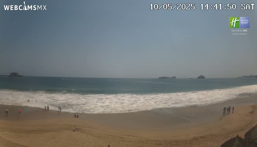 A sandy beach with a few people on it slopes down to a light-blue ocean with small waves under a clear sky.