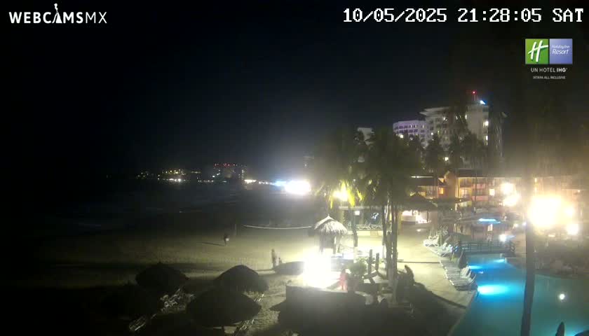 A nighttime beach scene shows a mostly empty sandy beach, palm trees, resort buildings with lights, and a swimming pool illuminated by artificial lighting.