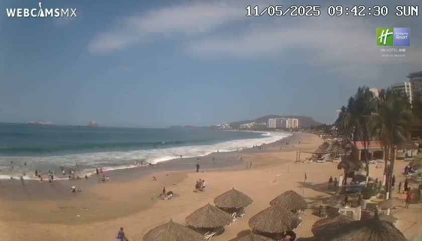 A sandy beach with a calm ocean and many people enjoying the sunny day,  with palm trees and buildings visible in the background.