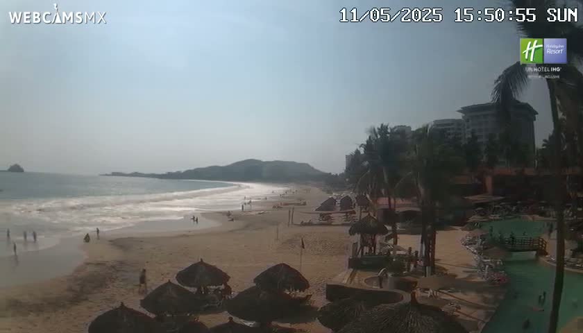 A sandy beach with calm waves, a few people, and thatch-roofed umbrellas stretches out under a hazy sky, with palm trees and buildings visible in the background.