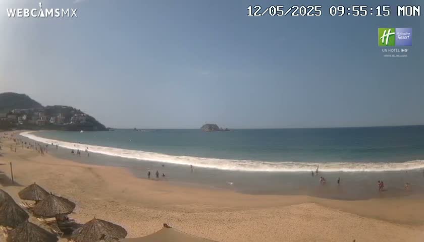A sandy beach with calm ocean waves stretches before a hillside of buildings under a clear, sunny sky.