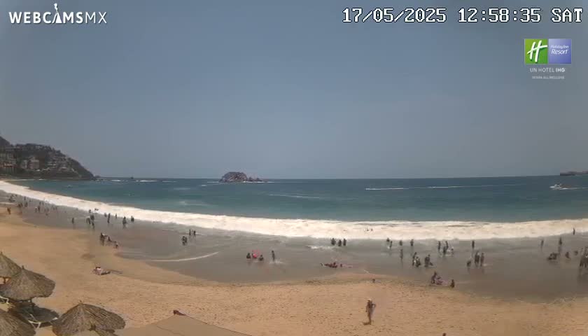 A sandy beach with many people on it and in the shallow water, under a clear, sunny sky, faces a calm ocean with small waves.