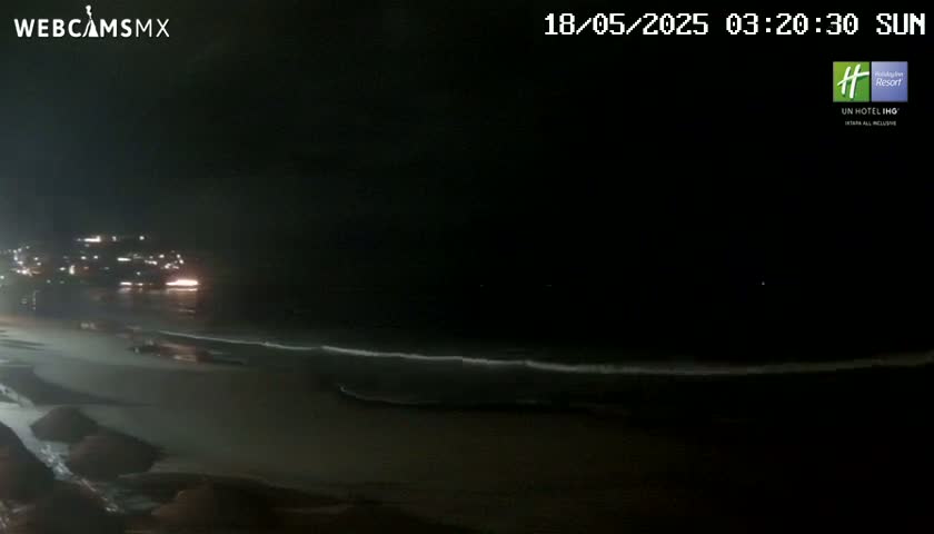 A nighttime beach scene shows dark water with gentle waves lapping a dimly lit shore under a cloudy sky.