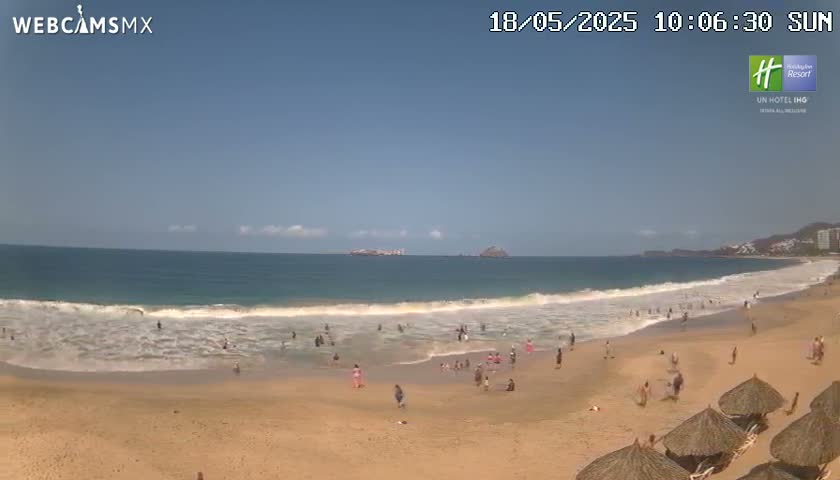 A sandy beach with many people swimming and relaxing under a mostly clear, sunny sky, with gentle waves rolling onto the shore.