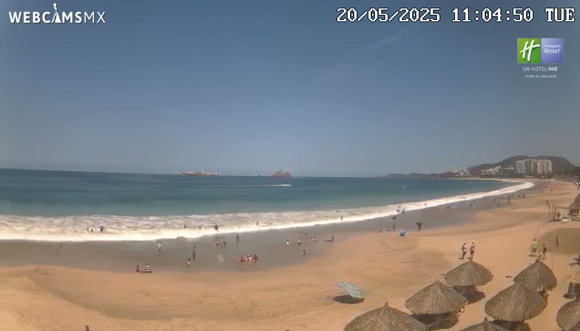 A sandy beach with calm waves and a few people on it stretches out to a clear blue ocean under a bright sunny sky, with buildings and hills visible in the distance.