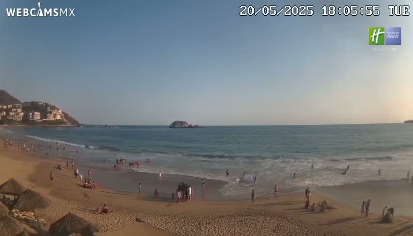 A sandy beach with calm waves and numerous people enjoying the late afternoon sun, backed by a hillside of buildings.