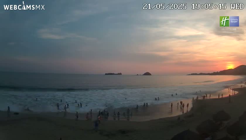 A beach scene at sunset shows people wading in the ocean waves as the sun sets over the horizon.