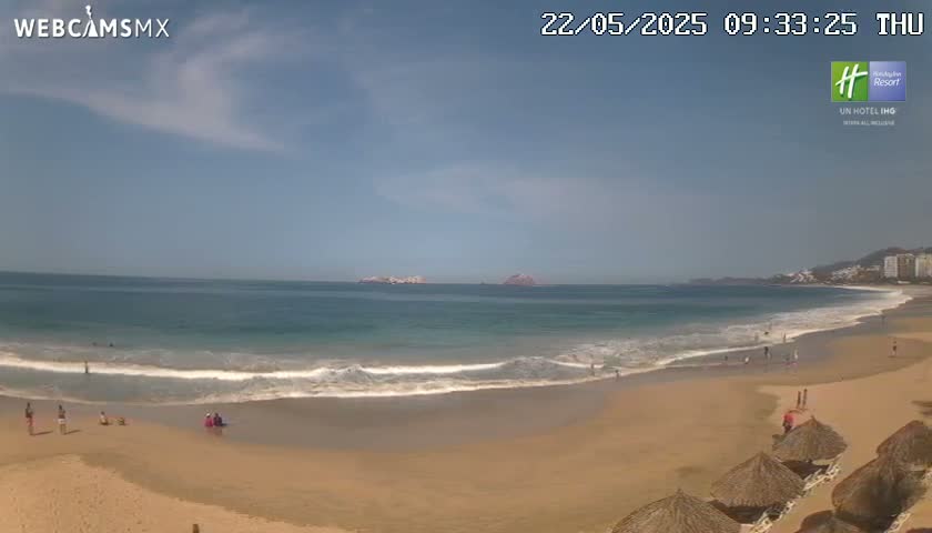A mostly sunny day at a beach with calm waves, light-brown sand, and several thatched umbrellas lining the shore.