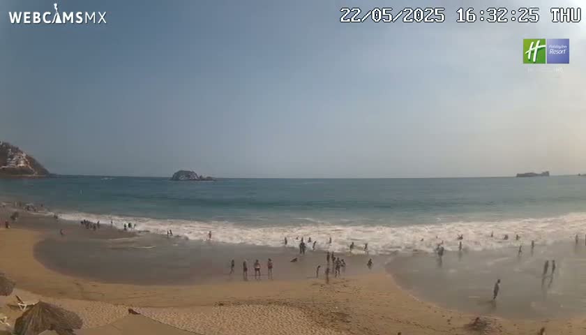 A sandy beach with people swimming and wading in the ocean under a clear, sunny sky.