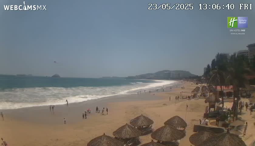 A sandy beach with a moderate surf under a clear, sunny sky has a scattering of people and several thatched umbrellas near some buildings.