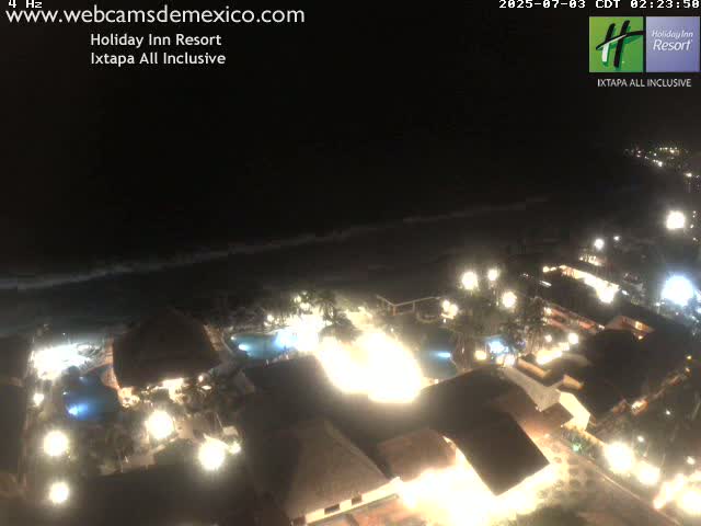 A brightly lit resort at night, situated near a dark ocean with waves visible under a dark sky.