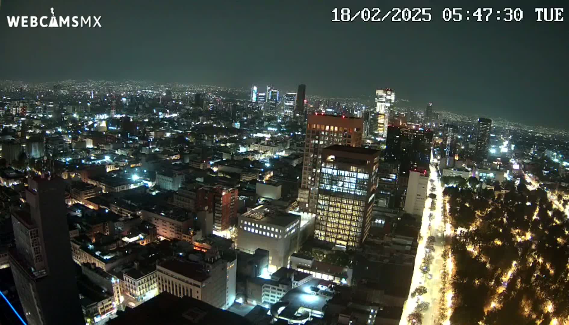 A nighttime aerial view of a city, showing numerous buildings illuminated with streetlights lining a wide avenue that cuts through a park.
