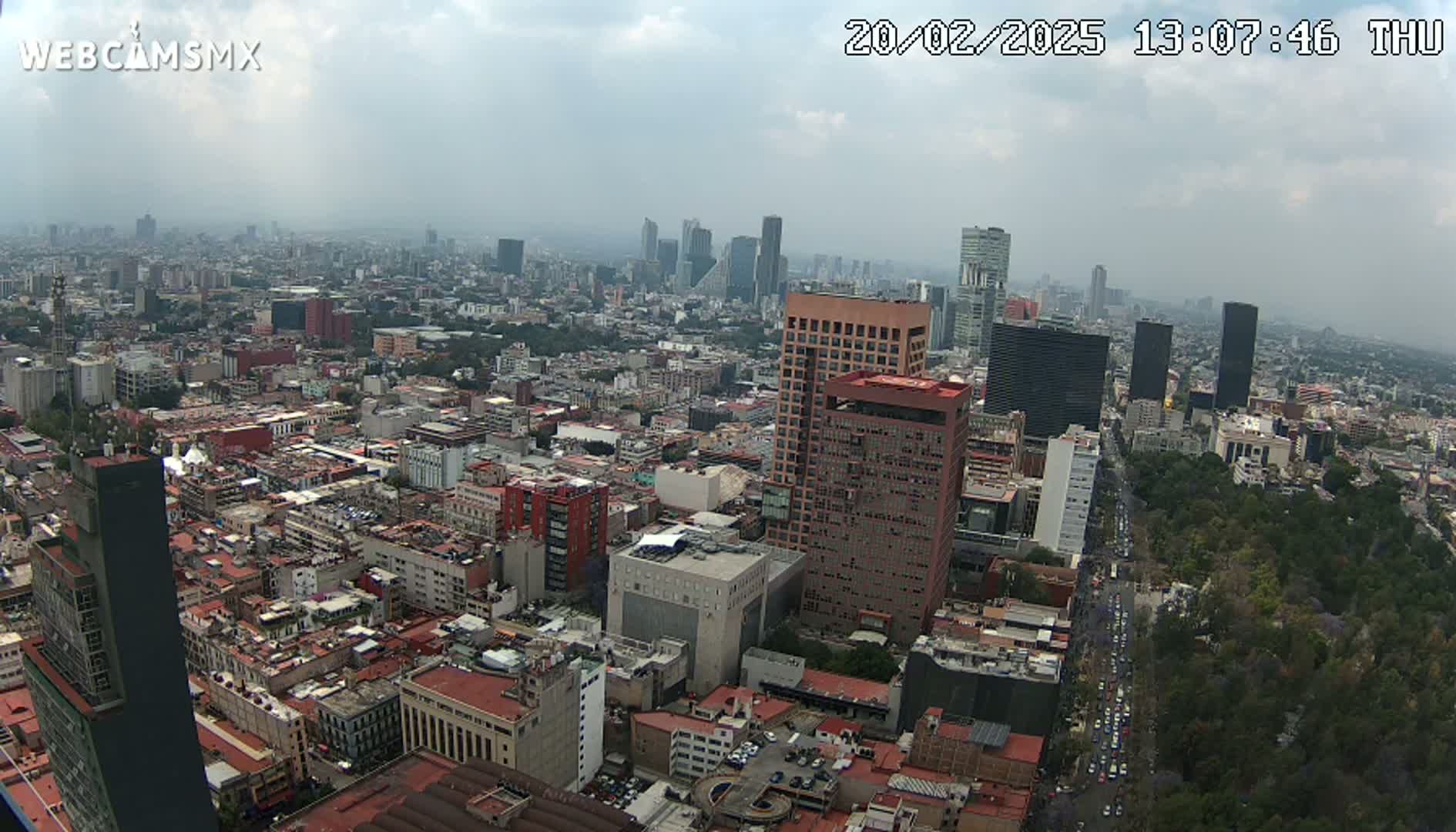 A hazy, high-angle view of a sprawling city with numerous buildings of varying heights, including skyscrapers and low-rise structures, a large park visible in the bottom right, and a wide avenue with traffic.
