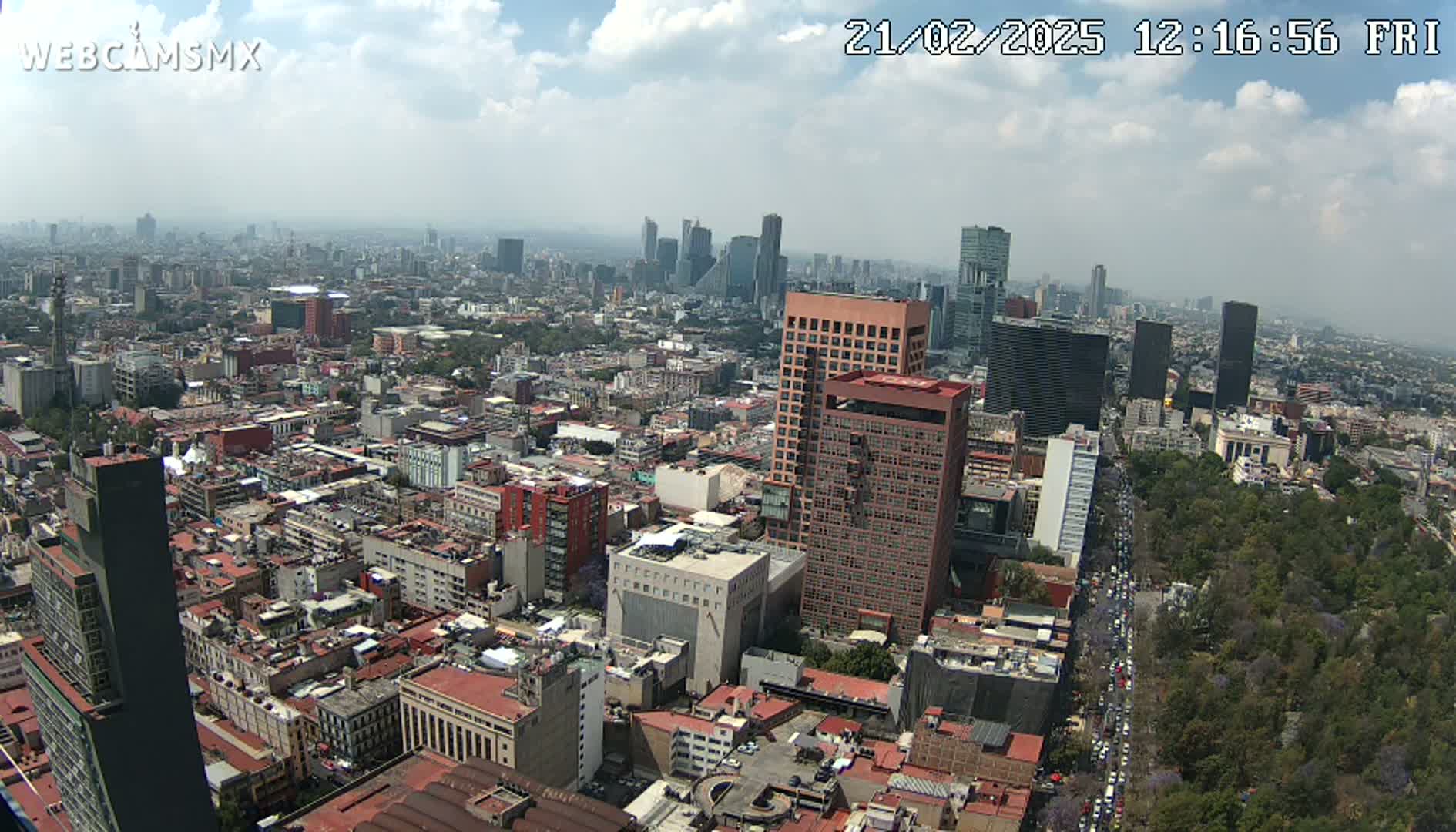 A hazy, partly cloudy day reveals a sprawling cityscape of Mexico City, with numerous buildings of varying heights and a large park visible in the lower right.