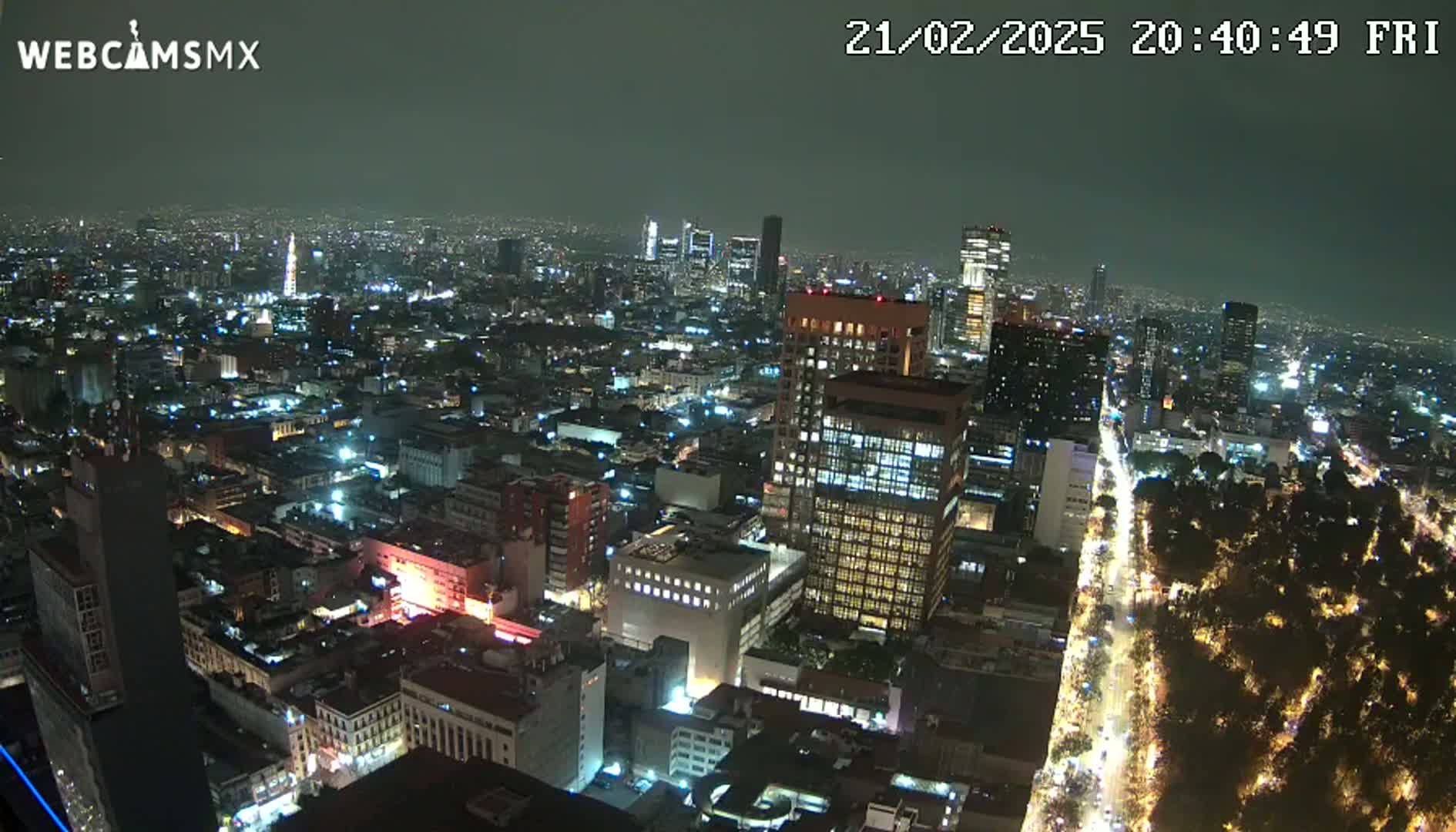 A nighttime aerial view of a large city with many illuminated buildings and streets, showing a park with lights along a major thoroughfare.