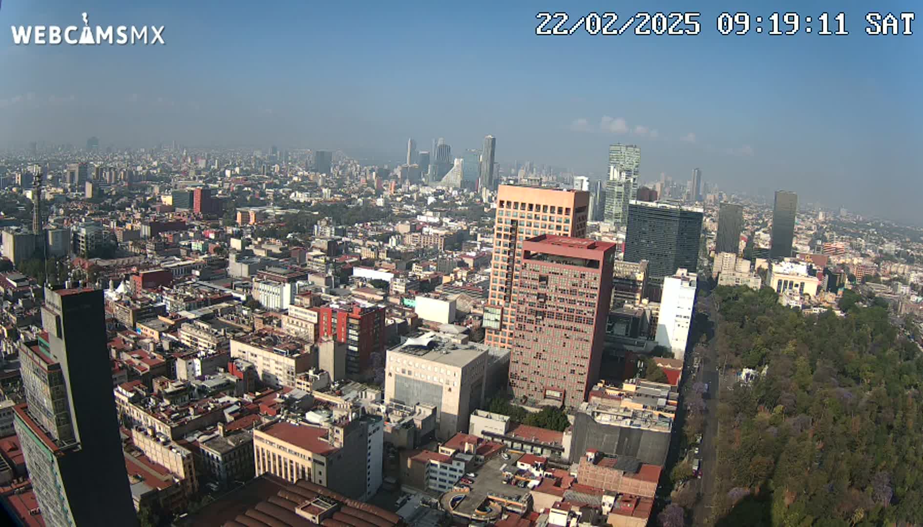 A hazy, sunny day view of a sprawling city with numerous buildings of varying heights, including several tall skyscrapers, and a large park area visible in the lower right.