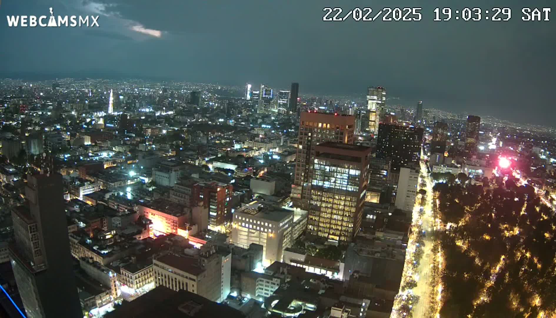A nighttime, high-angle view of a sprawling city illuminated by lights, with a partly cloudy sky.