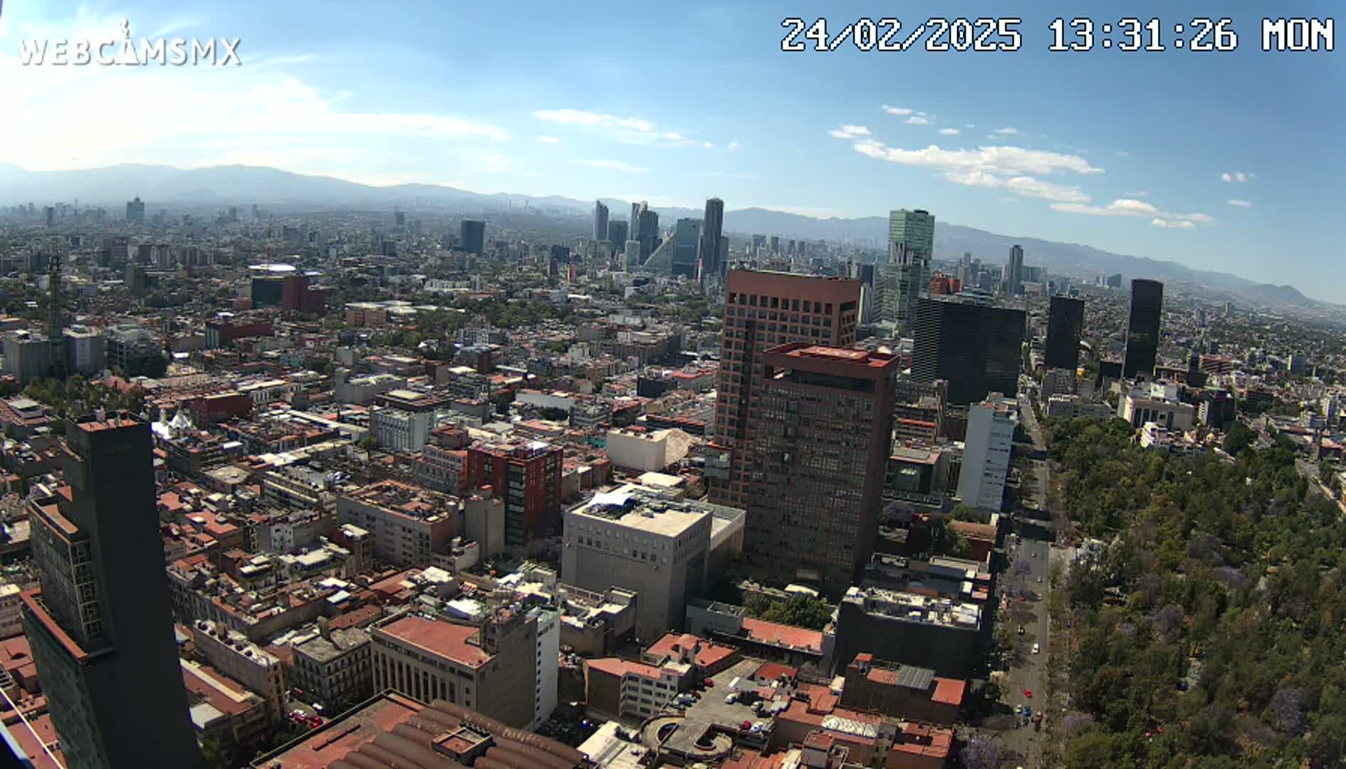 A high-angle, long shot on a partly cloudy day shows a sprawling city with numerous buildings of varying heights, some skyscrapers, and a large expanse of green parkland, all set against a backdrop of distant mountains.