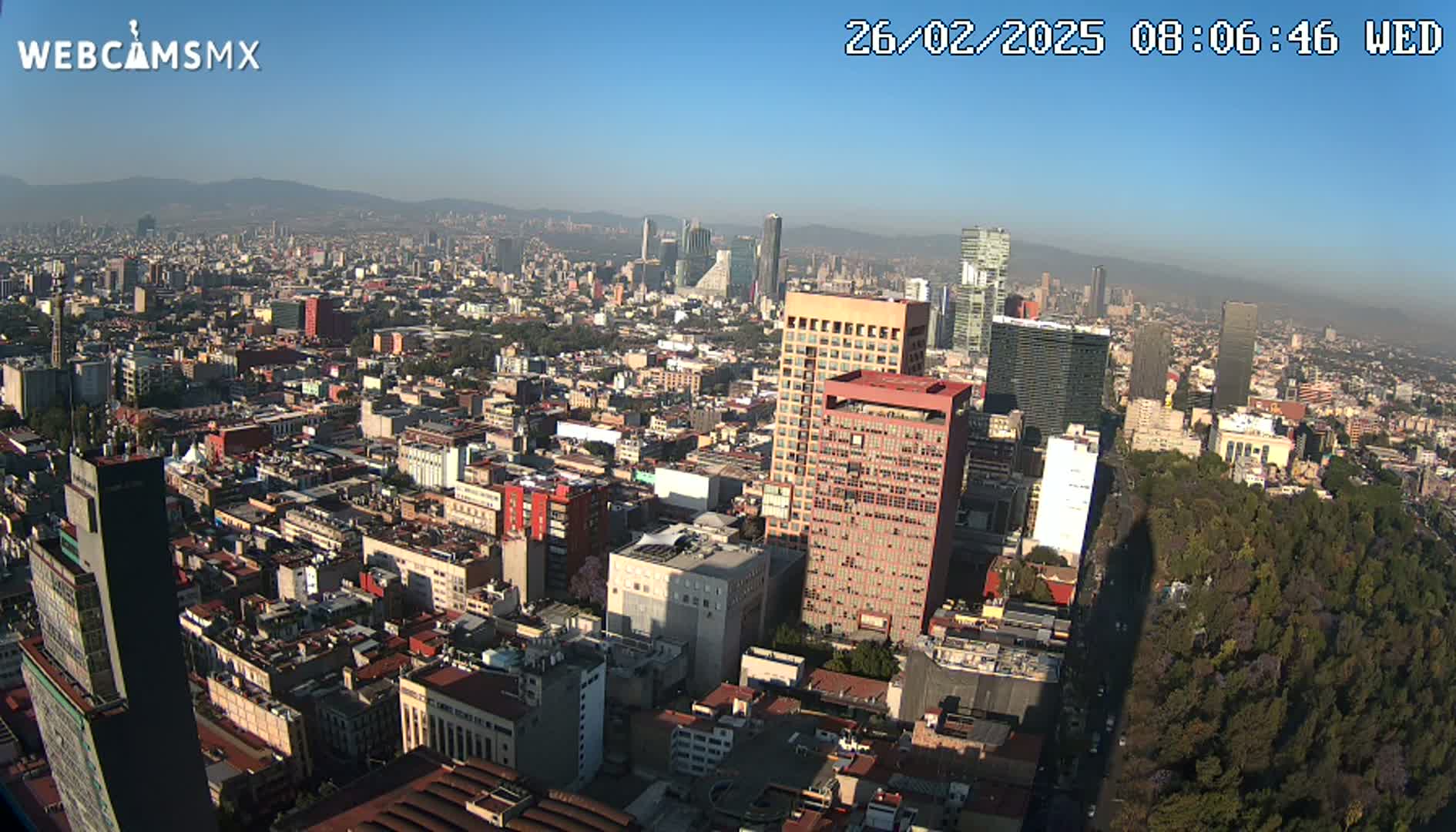 A high-angle, long shot shows a sprawling cityscape under a clear, sunny sky, with a large park visible in the lower right.