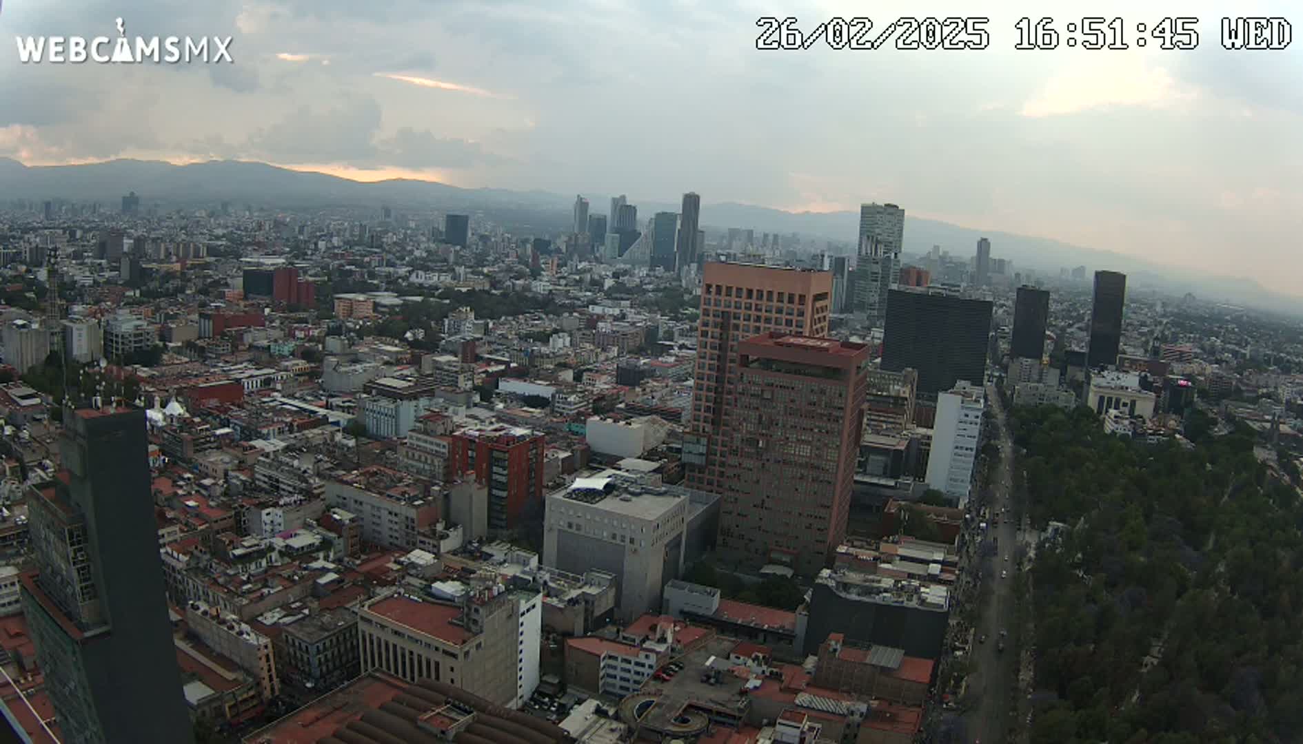 A high-angle, long shot of a sprawling city under a mostly cloudy sky, with a few taller buildings standing out against the dense urban landscape.