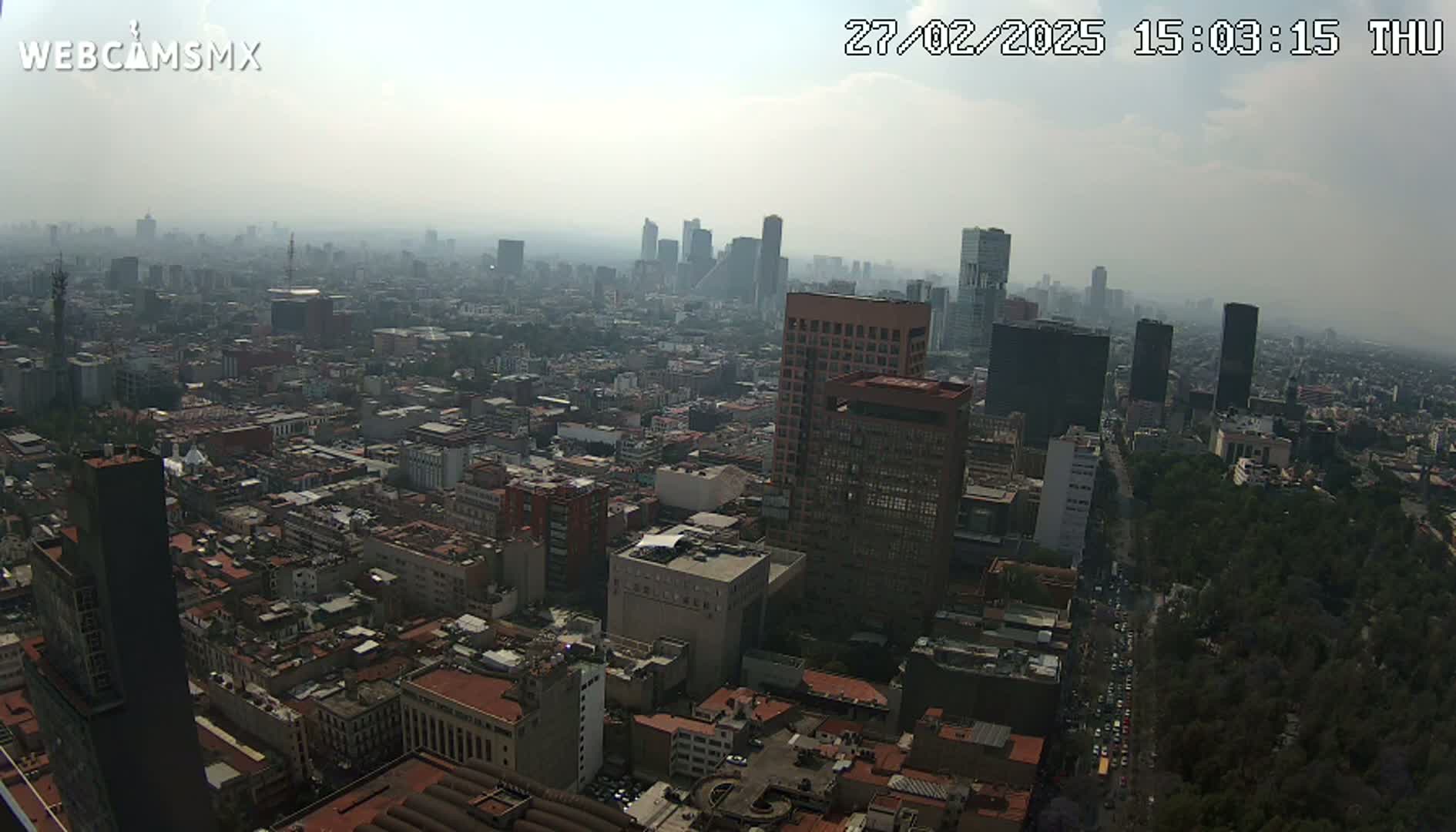 A hazy, high-angle view of a sprawling city, showing numerous buildings of varying heights, with a large park visible in the lower right.