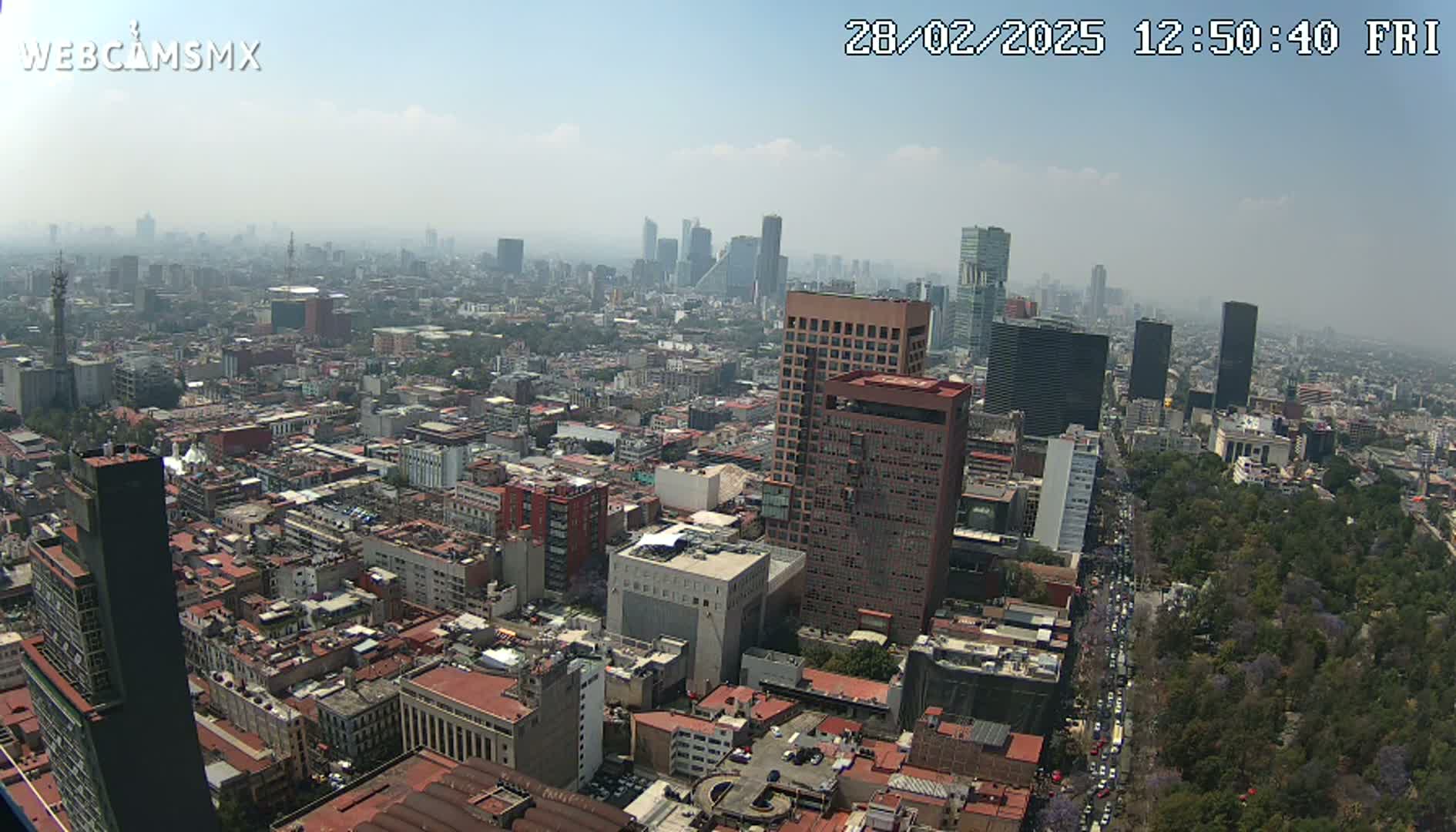 A hazy, high-angle view of a sprawling city with numerous buildings of varying heights, a large park, and a busy street visible in the lower right.