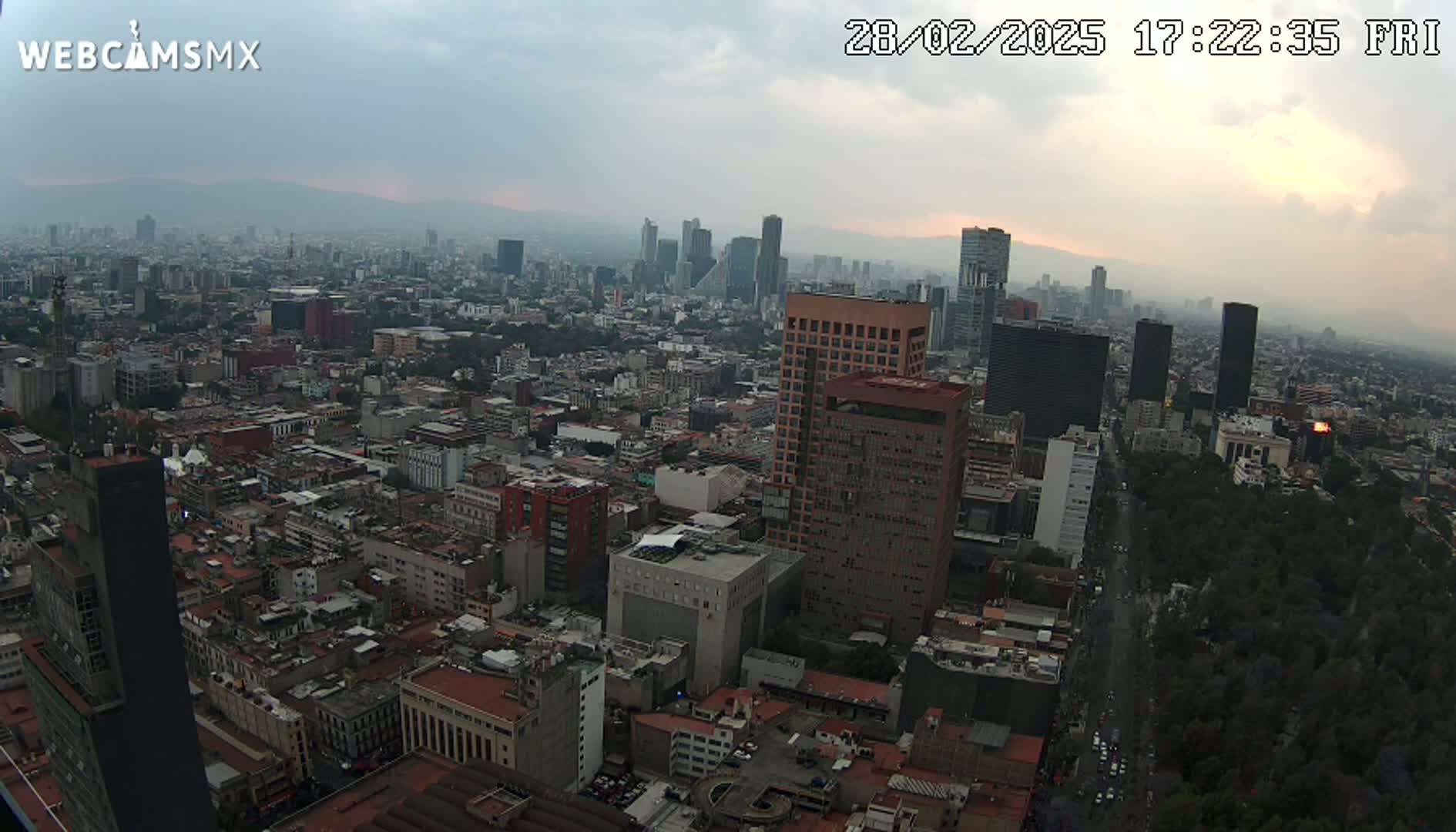 An expansive, hazy cityscape at sunset, featuring numerous buildings of varying heights and a long, tree-lined avenue.