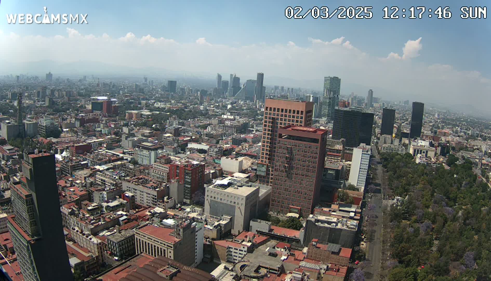 A high-angle, sunny view of a sprawling city with numerous buildings, including high-rises, under partly cloudy skies.