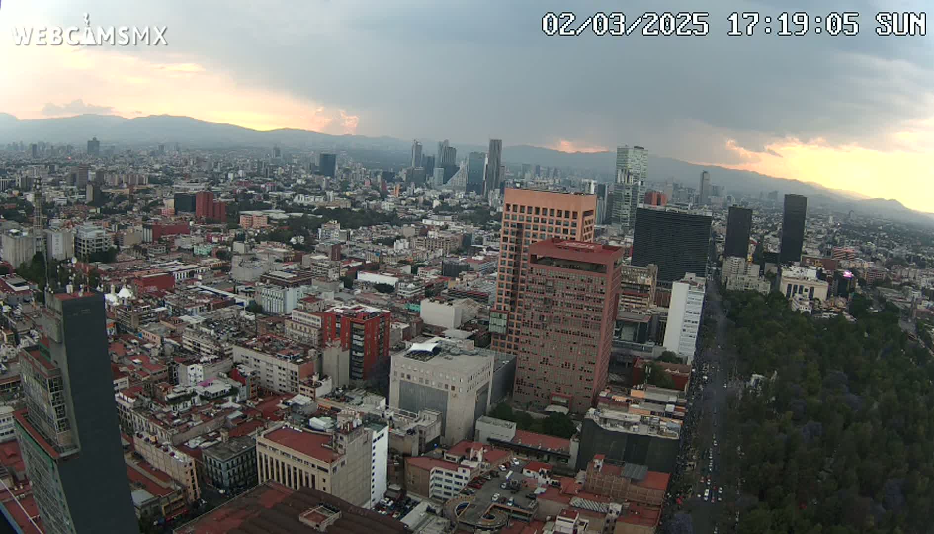 An aerial view of a sprawling city at sunset under partly cloudy skies, showing numerous buildings and a large park.