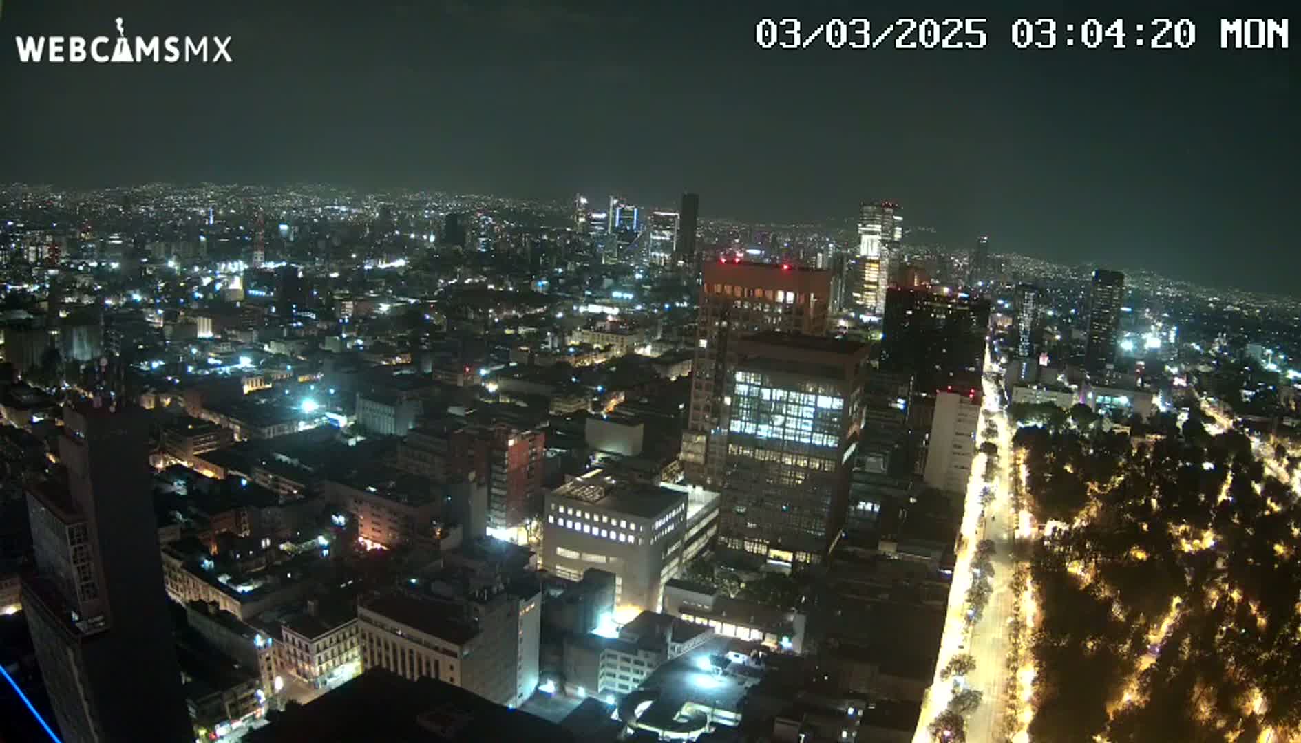 A nighttime aerial view of a city with brightly lit buildings and streets, and a tree-lined avenue.
