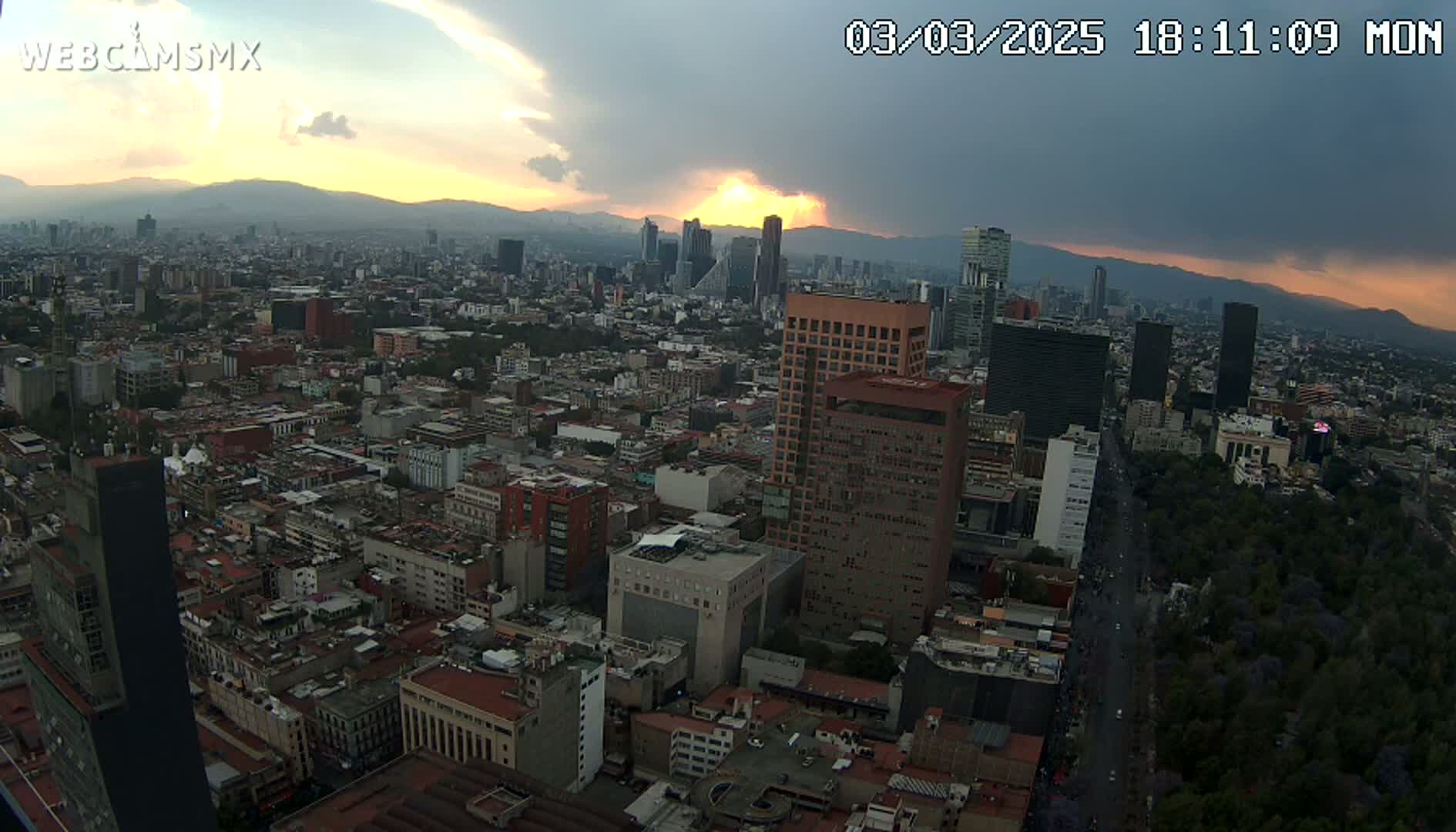 A sprawling cityscape at sunset, with partly cloudy skies and mountains in the distance.