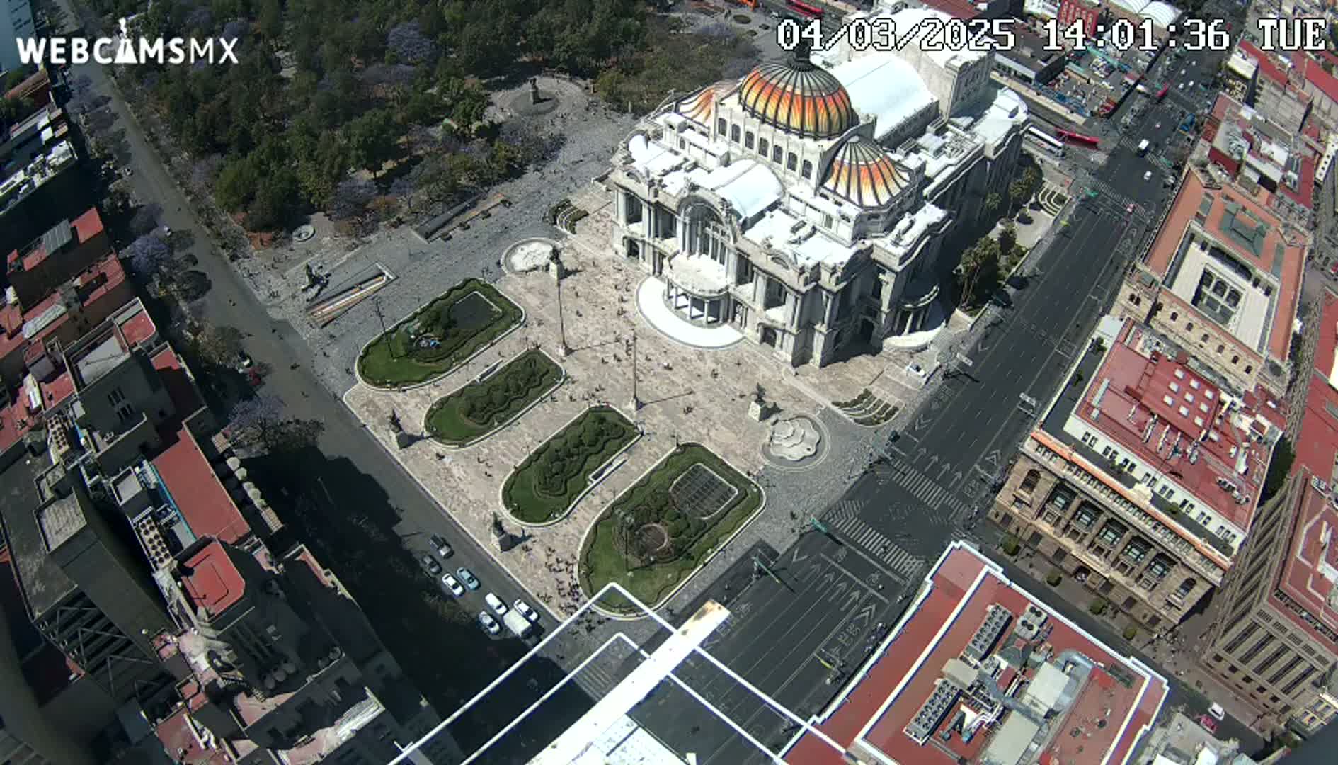 An aerial view of a city square with a large, ornate building, surrounding buildings, and a few vehicles on a sunny day.