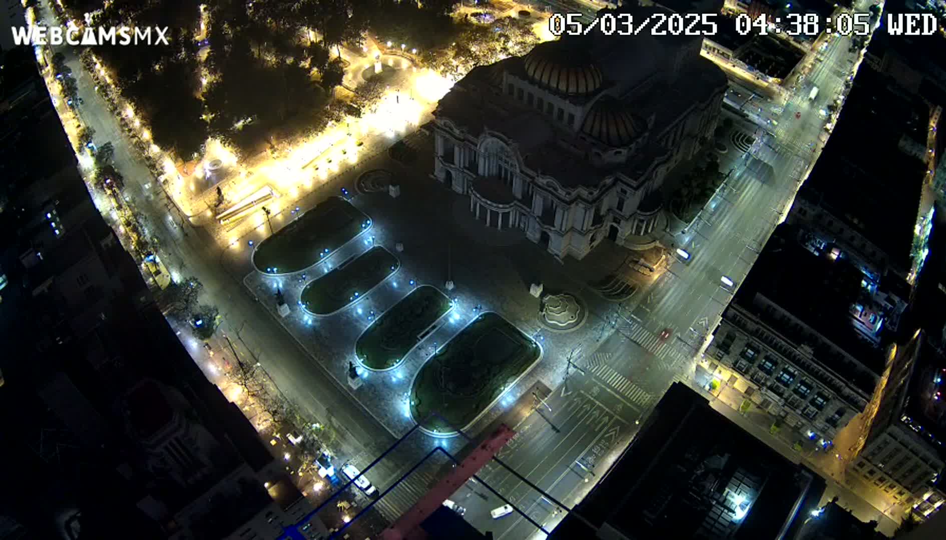 An aerial nighttime view of a city square with a large building, illuminated landscaping, and sparsely trafficked streets.