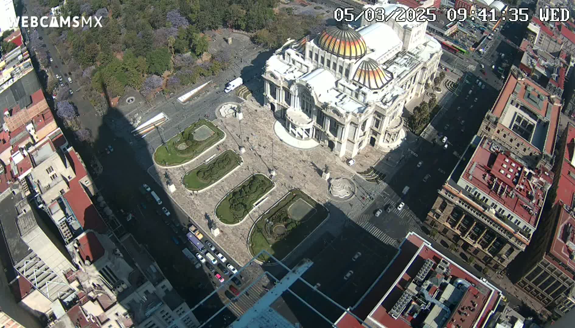 A high-angle, sunny view shows a large plaza with a palace-like building, surrounding buildings, and a long shadow cast from a tall structure, all under a clear sky.