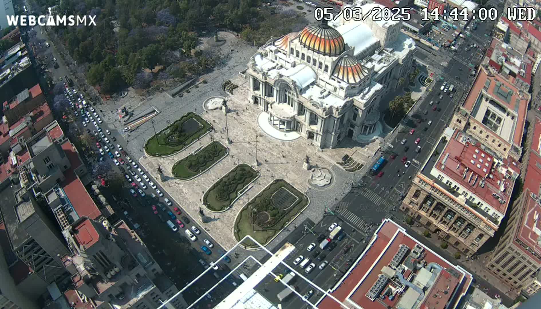 An aerial view shows a large building with a dome in a plaza, surrounded by city streets with cars and buildings, on a sunny day.