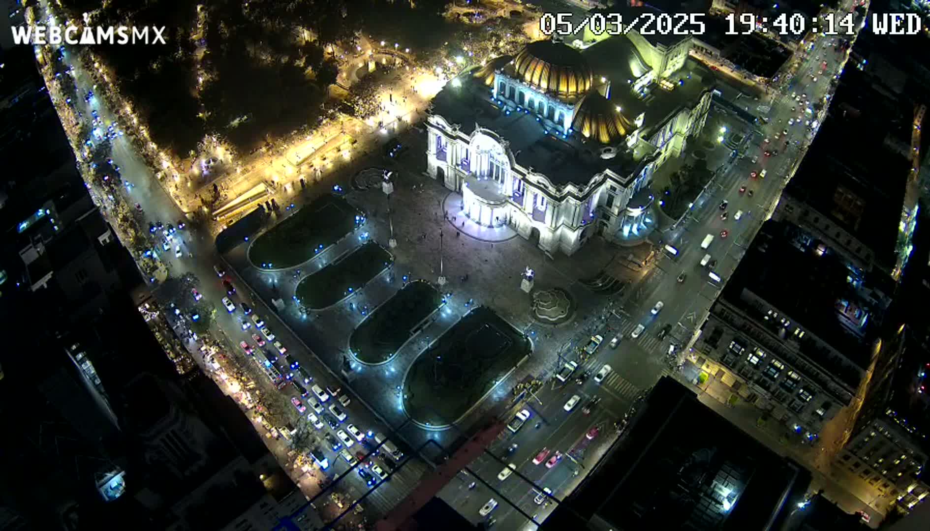 An aerial nighttime view shows a brightly lit palace-like building surrounded by streets with cars and a plaza with landscaped areas.