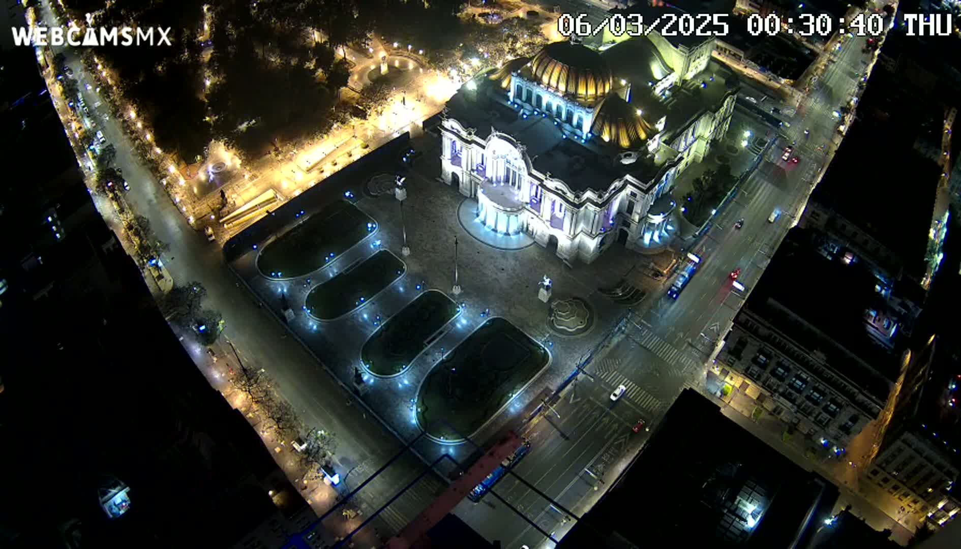 An aerial nighttime view shows a large, ornate building brightly lit, surrounded by a plaza with landscaping and streets with moving vehicles.