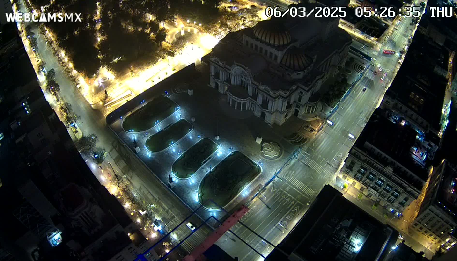 An aerial nighttime view of a city square with a large building, illuminated landscaping, and sparsely trafficked streets.