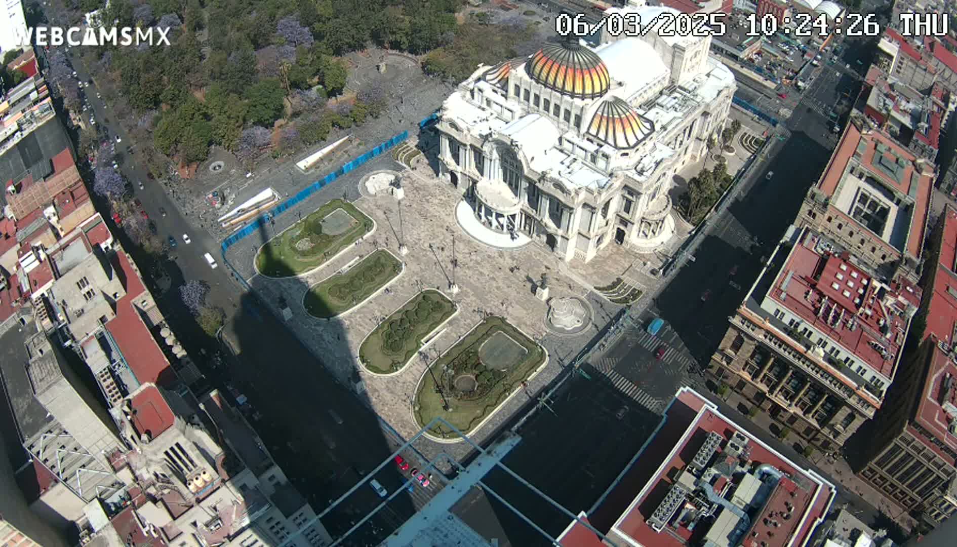 An aerial view shows a large, ornate building in a city square surrounded by other buildings and a park-like area on a sunny day.