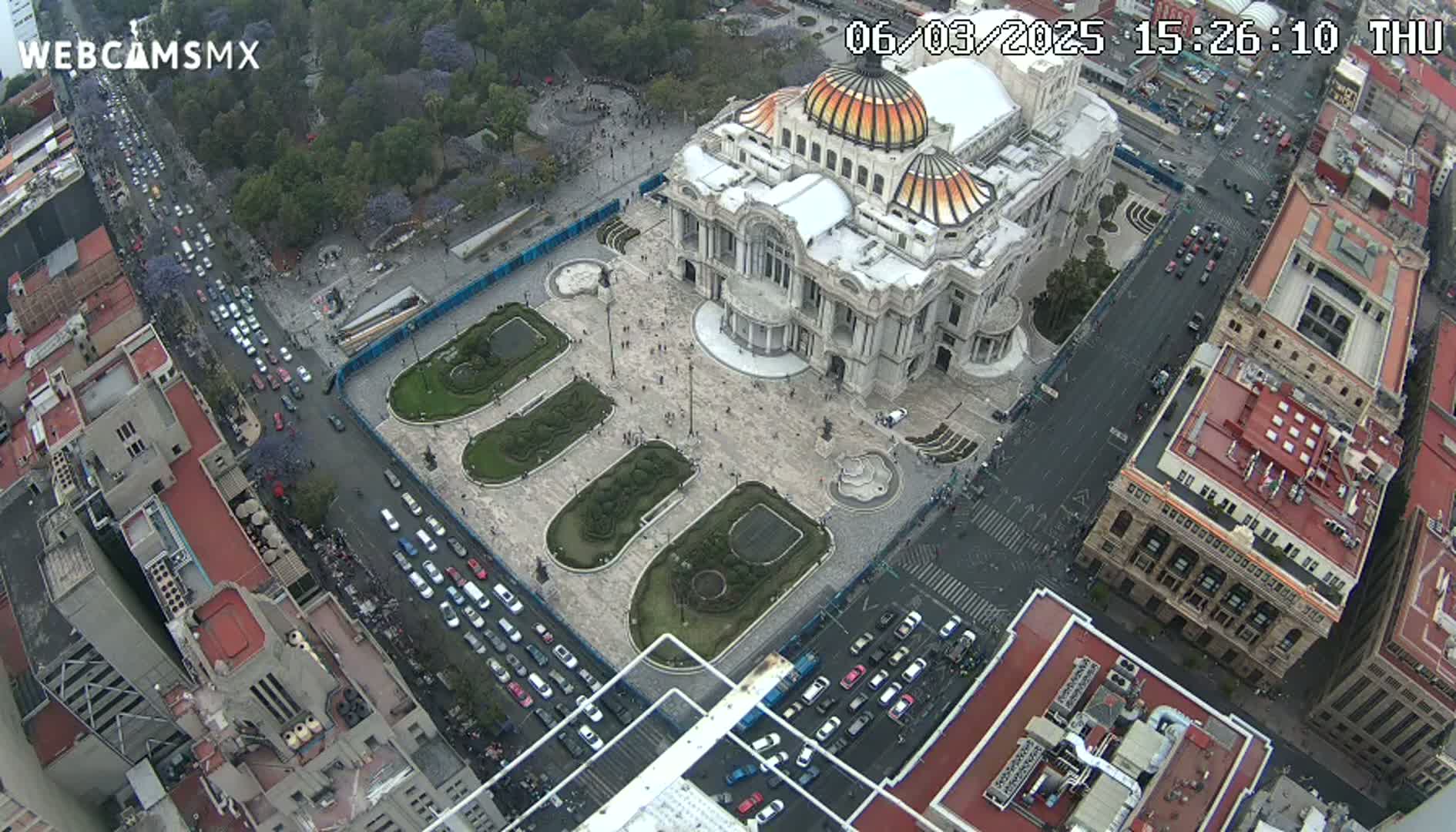 An aerial view shows a large, ornate building in a city square surrounded by streets with parked cars and green landscaping under a partly cloudy sky.